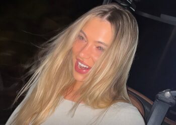 Smiling woman with long blonde hair and light makeup wearing a light top in a car at night with a seatbelt visible