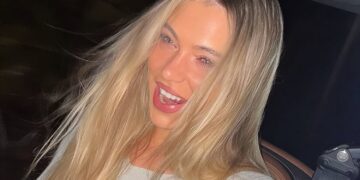 Smiling woman with long blonde hair and light makeup, wearing a light top in a car at night, with a seatbelt visible.