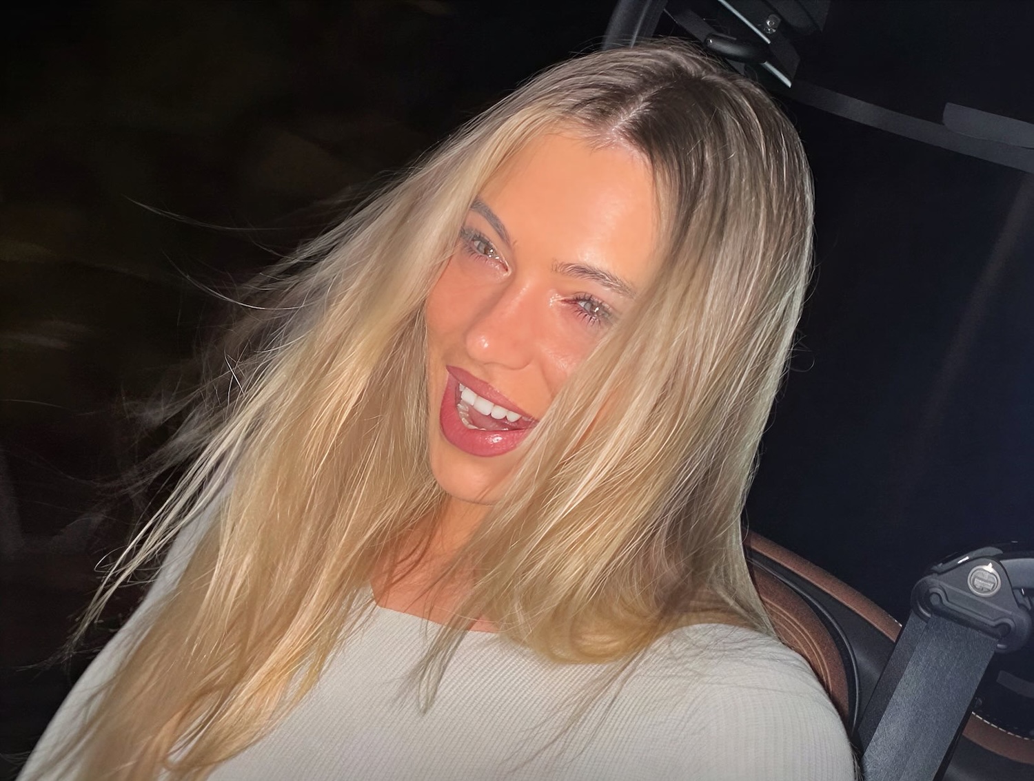 Smiling woman with long blonde hair and light makeup wearing a light top in a car at night with a seatbelt visible
