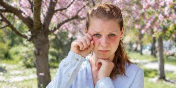 Woman in a light blue shirt rubbing her eye while standing under pink blossoming trees in a sunny park.