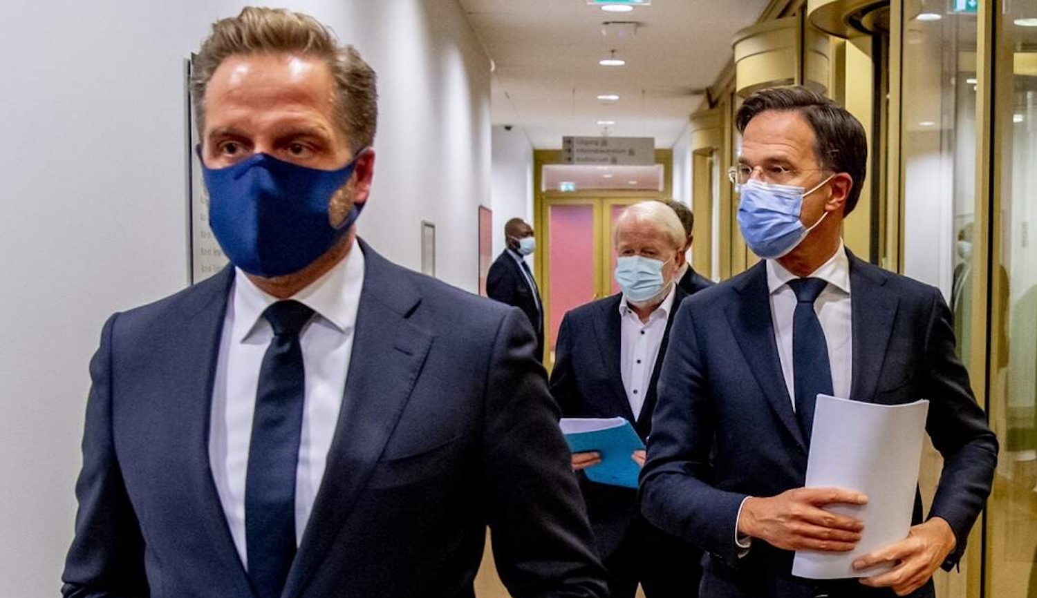 Three men in dark suits wearing protective face masks walk down a bright hallway one holds a blue folder and another holds a stack of papers
