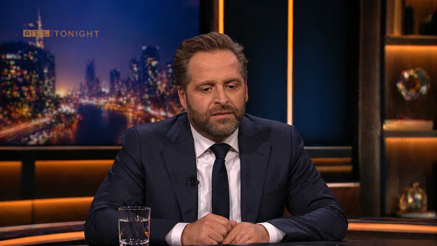 Man in a navy suit and tie sits at a desk on a television talk show set with a city skyline backdrop and a glass of water in front of him