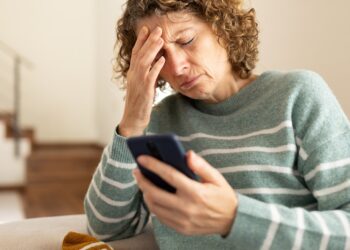 Woman sitting on a couch, holding her forehead in frustration while looking at a smartphone in her hand at home.