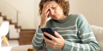 Woman sitting on a couch, holding her forehead in frustration while looking at a smartphone in her hand at home.