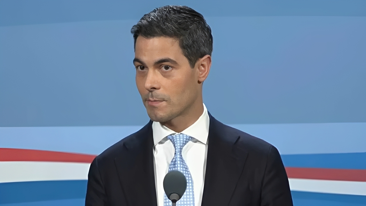 Man in a dark suit and light blue tie speaks at a microphone against a blue stage backdrop to a formal audience