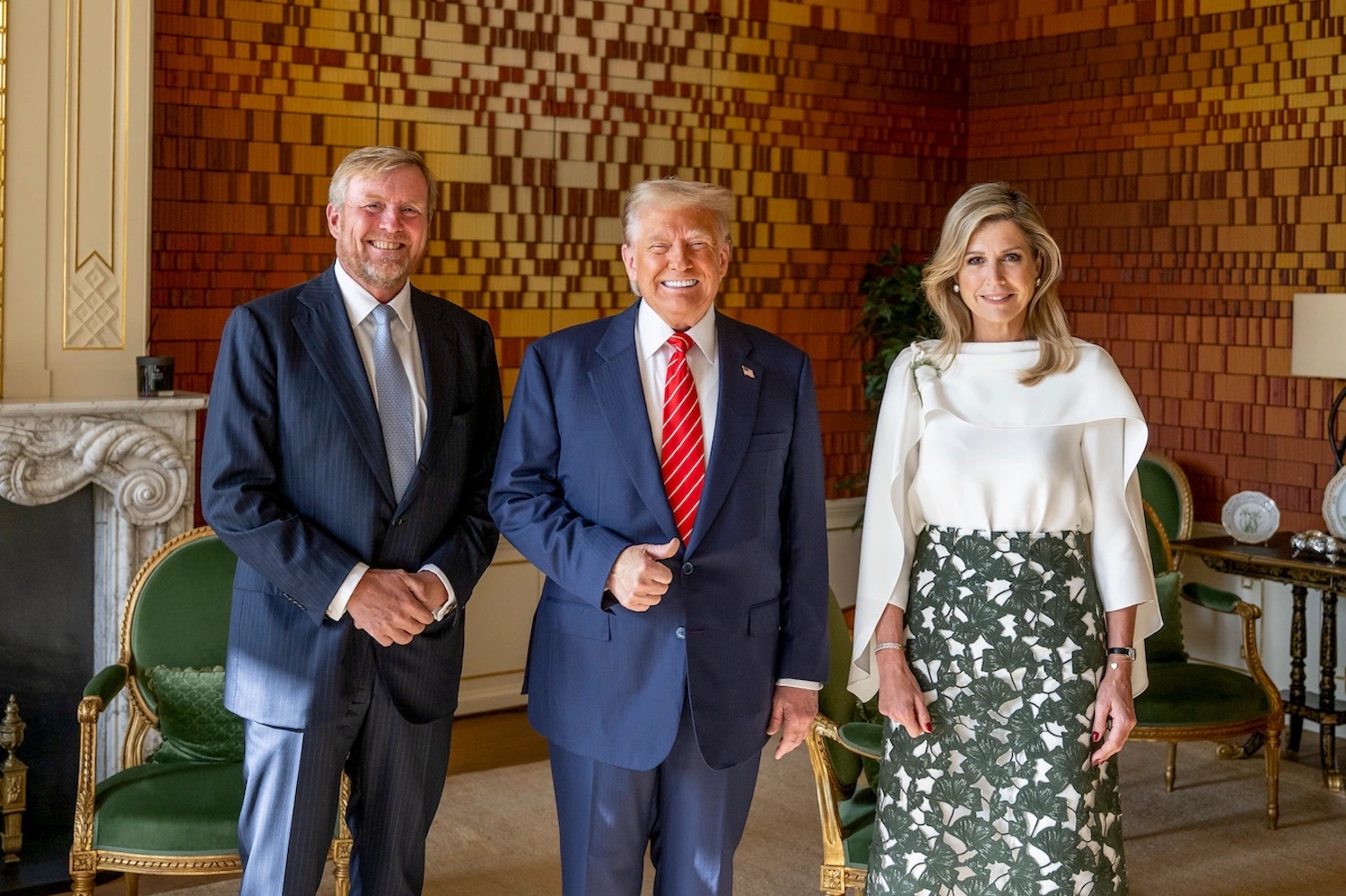 Three adults in formal attire pose in an ornate room center man in navy suit with red tie woman in white top and green floral skirt to his right man in dark suit to his left all smiling