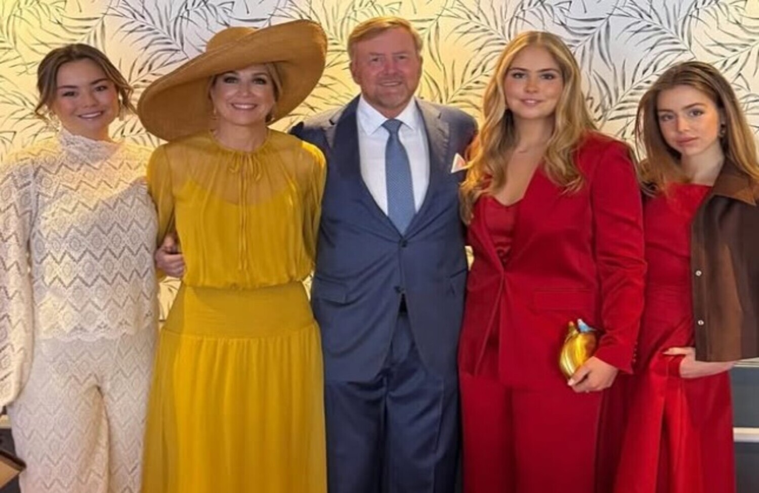 Five adults posing in formal attire at a stylish event woman in a white lace dress woman in mustard yellow with a wide hat man in a suit and two women in red outfits