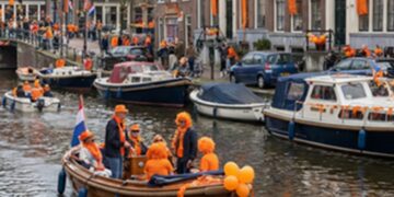People in orange outfits on a wooden boat in a canal parade with orange decorations and houses in the background