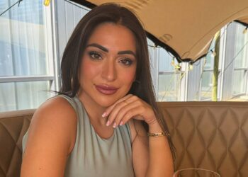 Portrait of a woman with long dark hair wearing a light blue sleeveless top resting her chin on her hand in a tan quilted restaurant booth