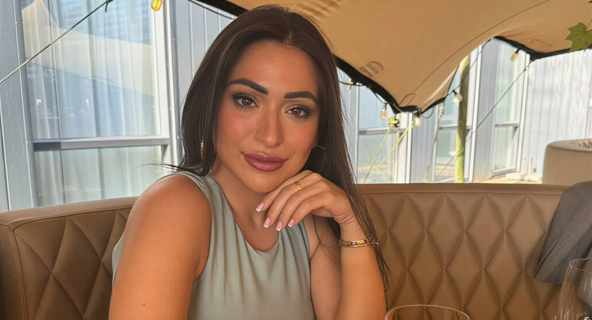 - Info Vandaag Portrait of a woman with long dark hair wearing a light blue sleeveless top resting her chin on her hand in a tan quilted restaurant booth