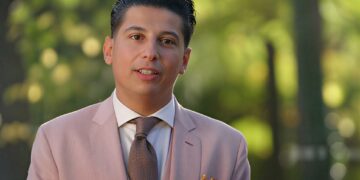 Portrait of a man in a light pink suit with a brown tie and purple boutonniere, standing outdoors with a blurred green background.