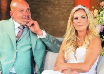 Bride and groom seated together at a wedding the groom in a teal suit and the bride in a white gown with a tiara with colorful flowers behind them