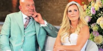 Bride and groom seated together at a wedding, the groom in a teal suit and the bride in a white gown with a tiara, with colorful flowers behind them.