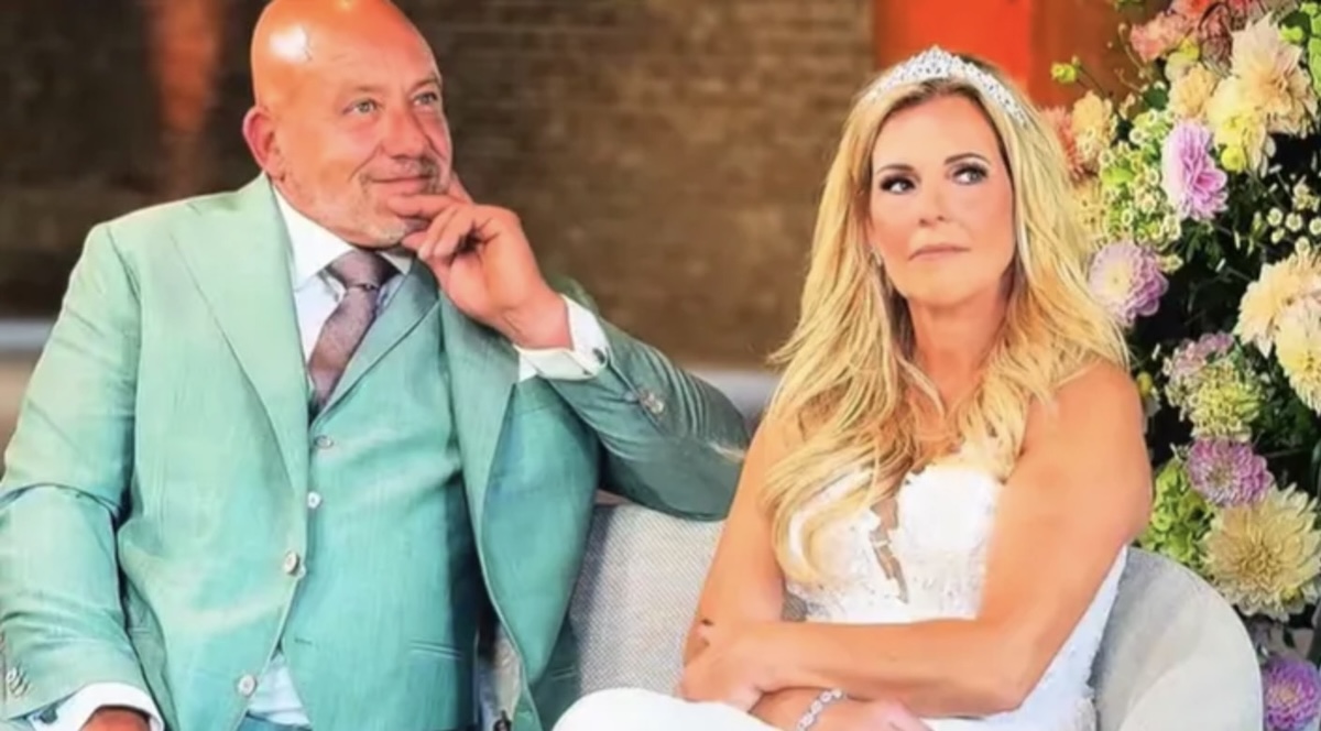 Bride and groom seated together at a wedding the groom in a teal suit and the bride in a white gown with a tiara with colorful flowers behind them
