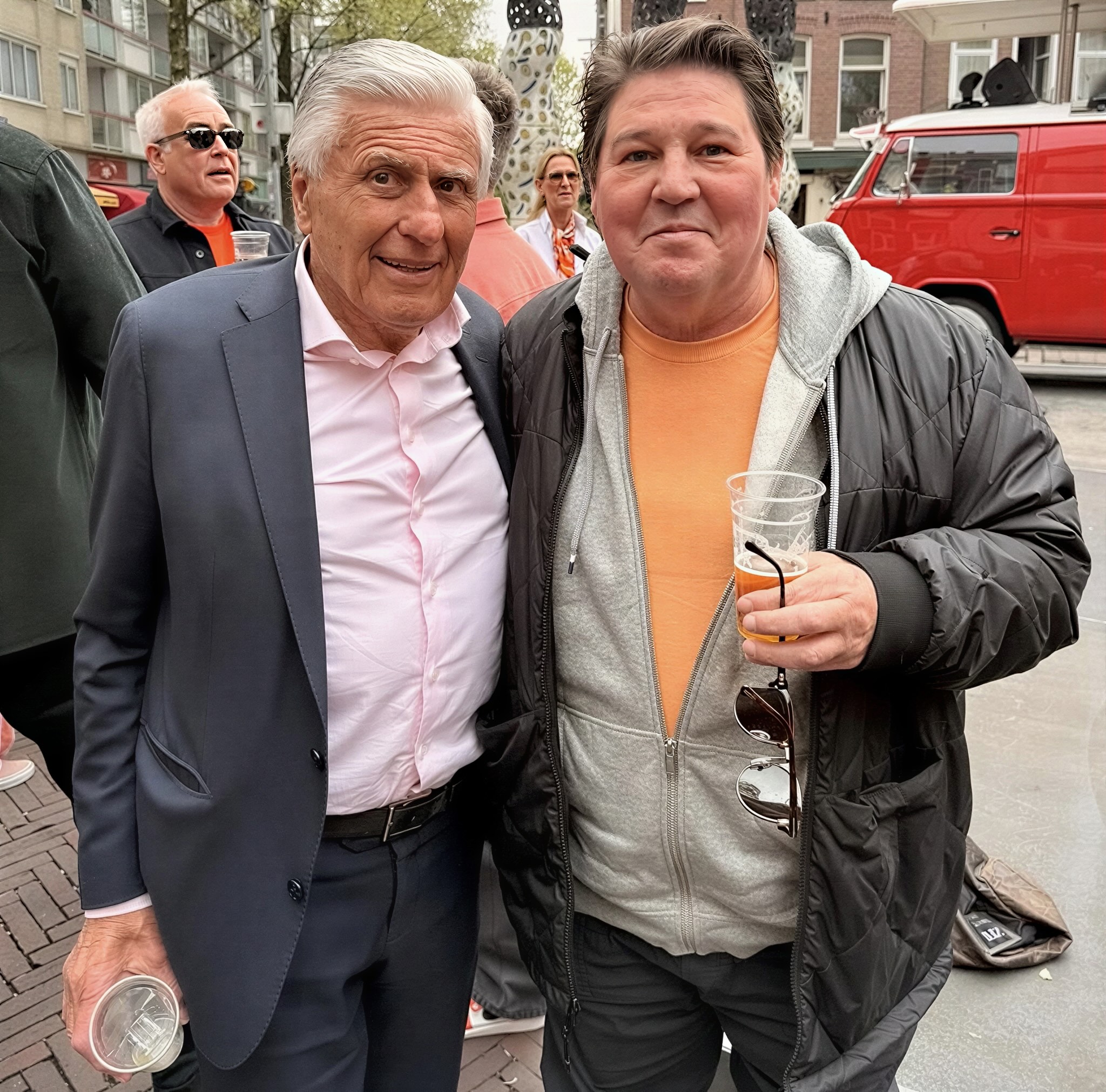 Two men posing at an outdoor city event an older man in a dark suit and pink shirt beside a younger man in a grey hoodie and orange shirt both holding plastic cups of beer