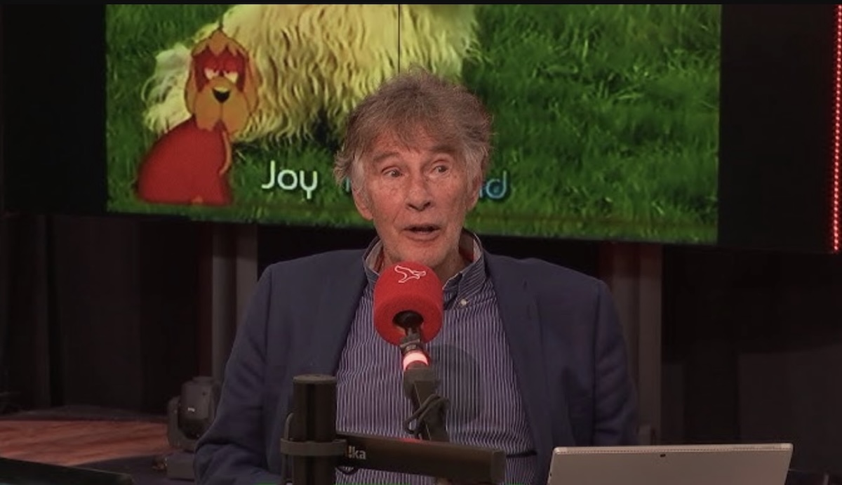 Older man in a blue blazer speaks into a red microphone at a studio desk with a screen showing a cartoon lion and 'joy' behind him