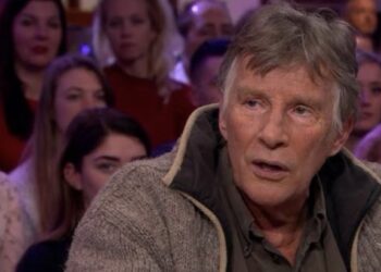 Older man with gray hair and a chunky beige cardigan sits in the foreground of a television talk show set with an audience visible in the blurred background