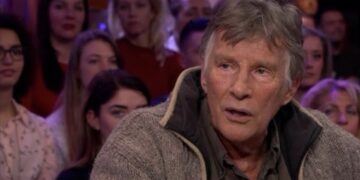 Older man with gray hair and a chunky beige cardigan sits in the foreground of a television talk show set, with an audience visible in the blurred background.