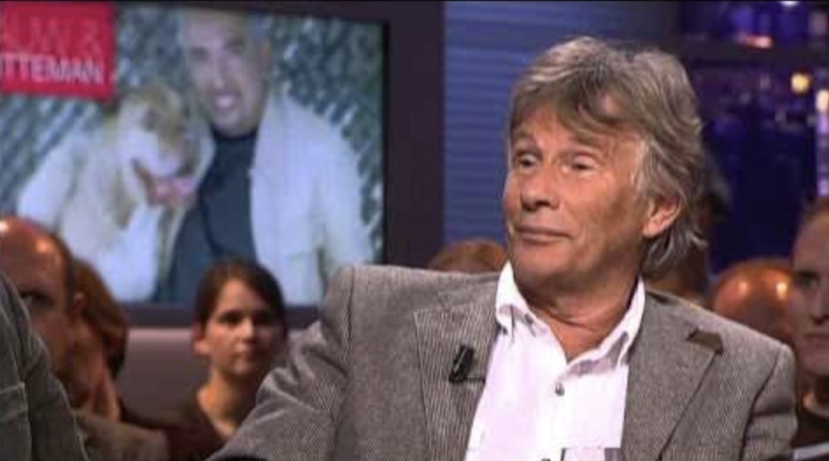 A gray haired man in a light blazer and white shirt sits on a tv talk show set with audience behind him and a large screen displaying a blurred image