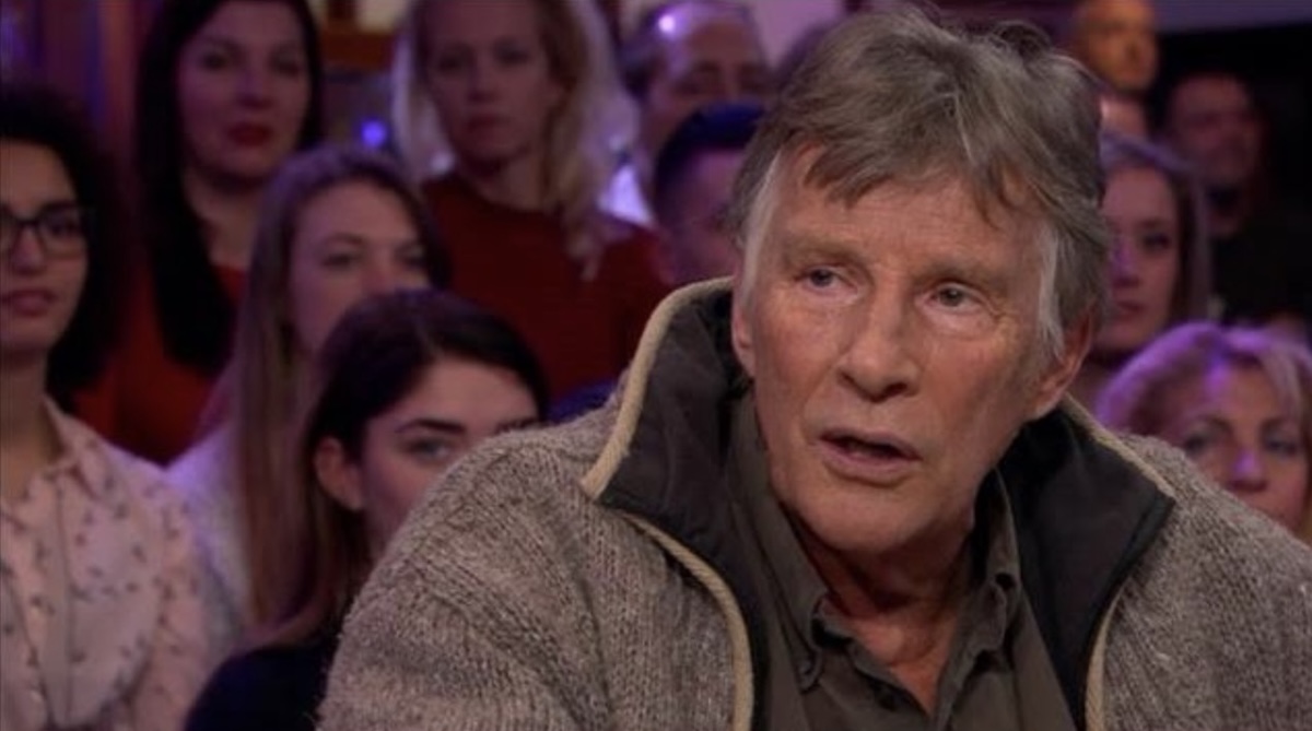 Older man with gray hair and a chunky beige cardigan sits in the foreground of a television talk show set with an audience visible in the blurred background
