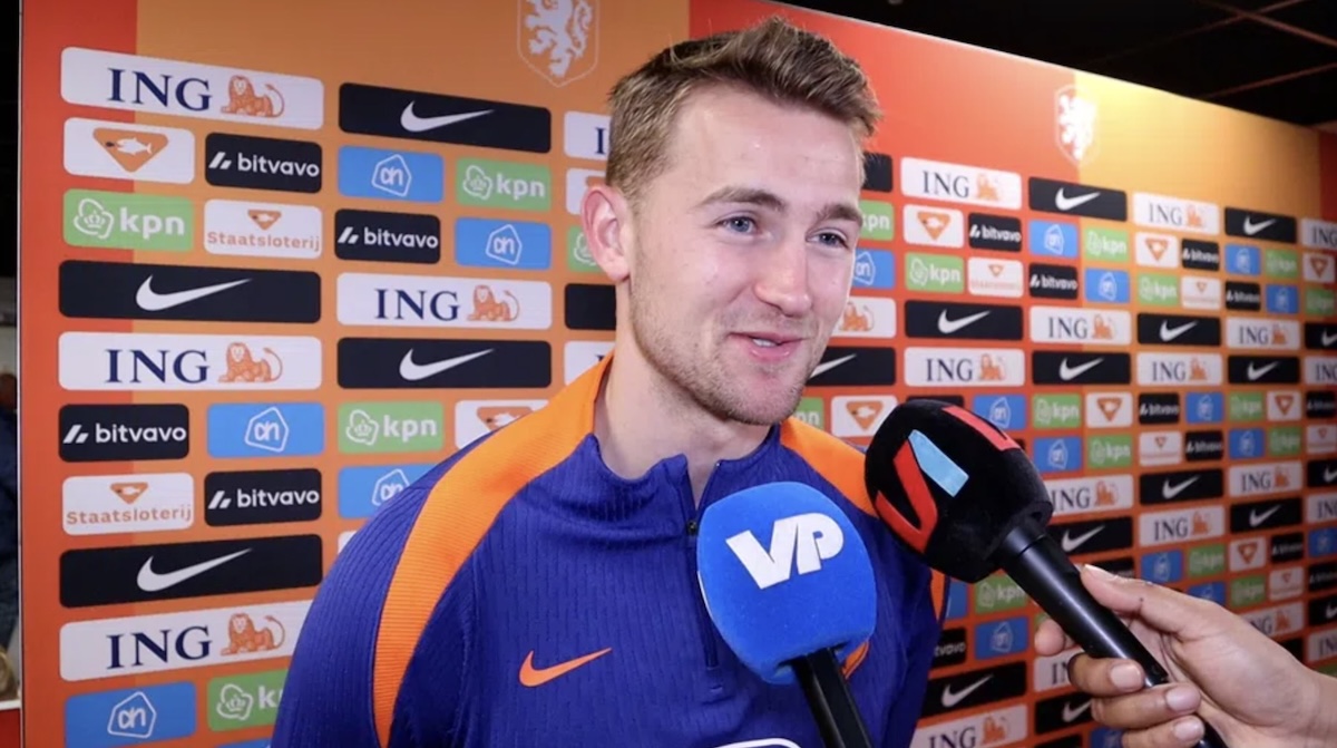 Male footballer in a blue and orange jersey giving a post match interview microphones in front of sponsor filled backdrop