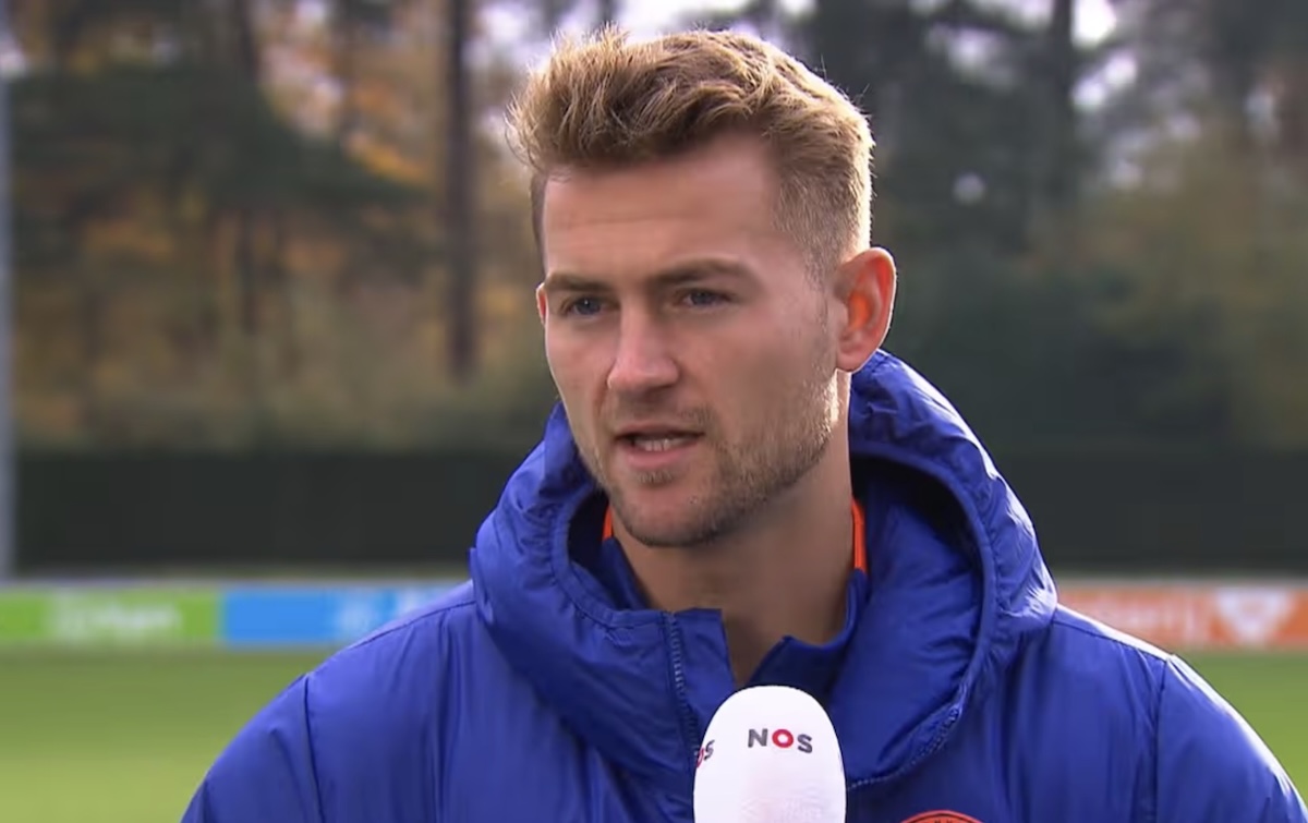 Man in a blue padded jacket is interviewed on a soccer training ground a nos mic held up to him