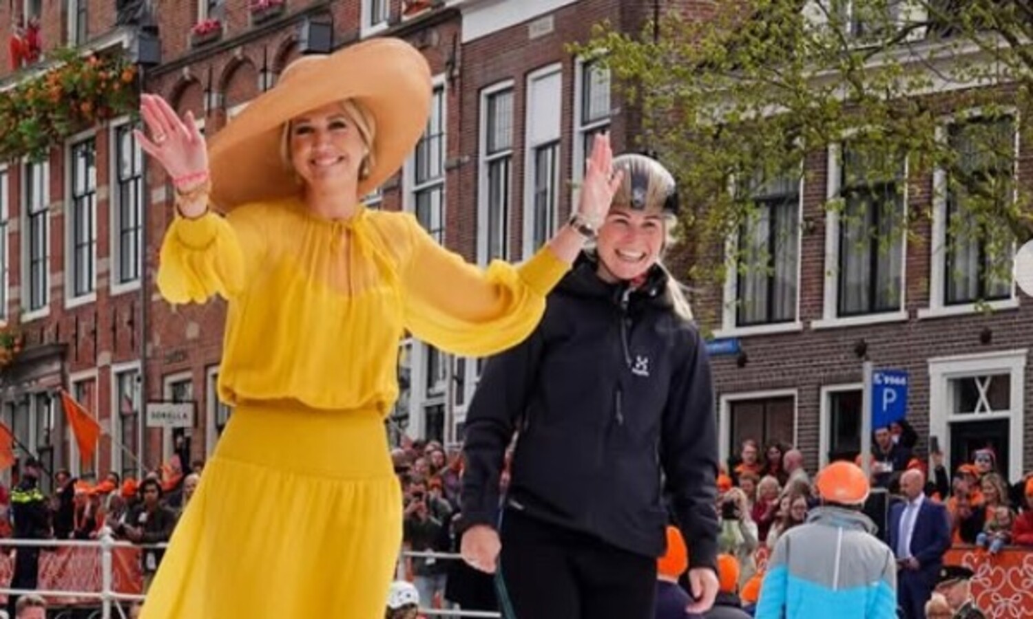 Two women cheerfully wave from a parade float one wears a bright yellow dress and wide brim hat the other a black jacket beside her