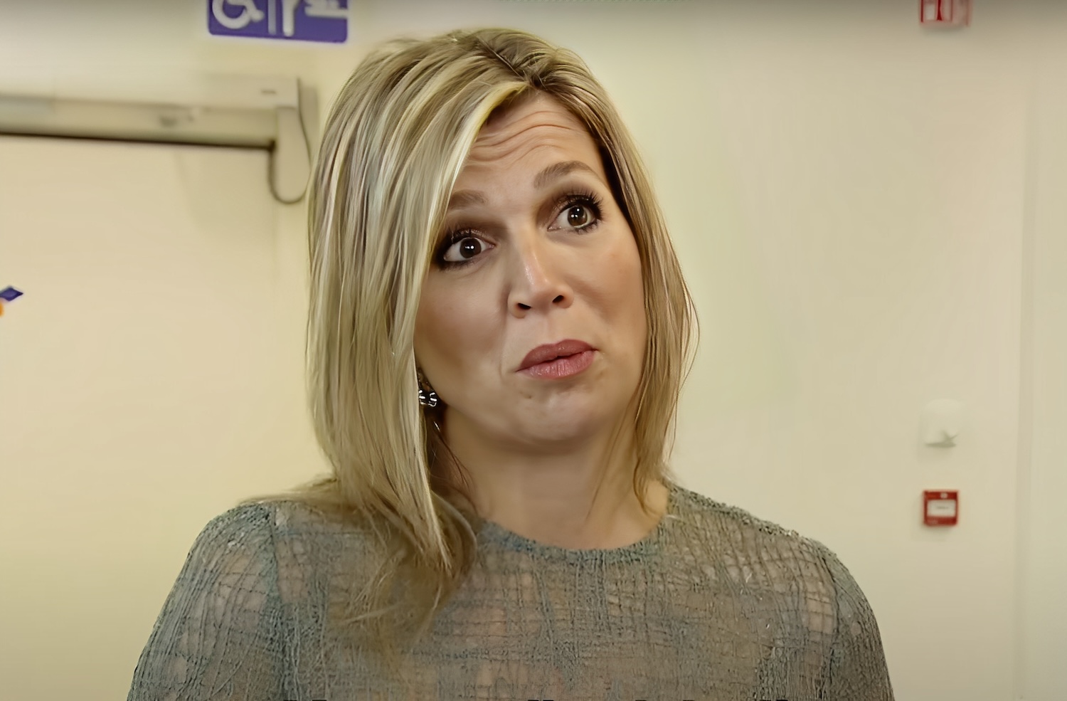 Woman in a gray knit top looks surprised or puzzled indoors near a hallway with a door behind her