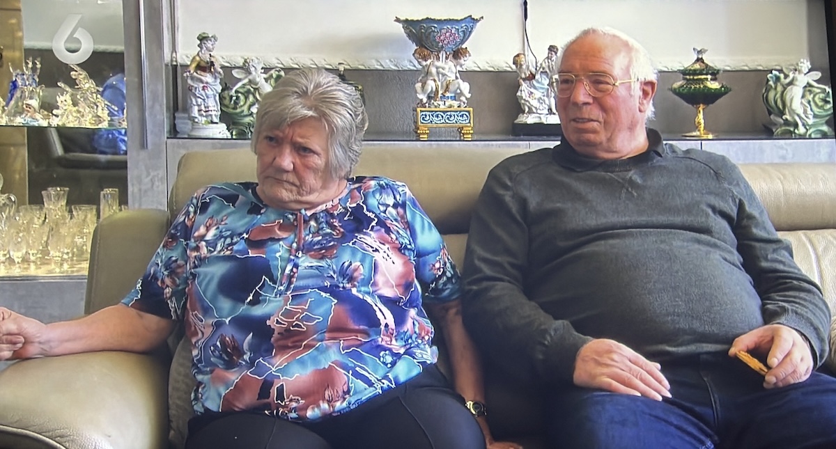 Couple seated on a beige sofa in a living room the woman in a colorful blue floral blouse and the man in a dark sweater holding a snack