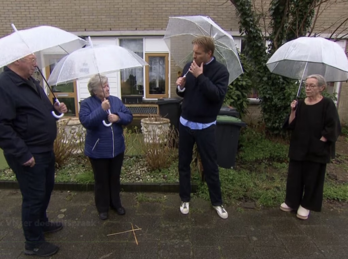 Five adults stand in a front yard circle under clear umbrellas in light rain with one man in the center smoking while talking to the group and others listening