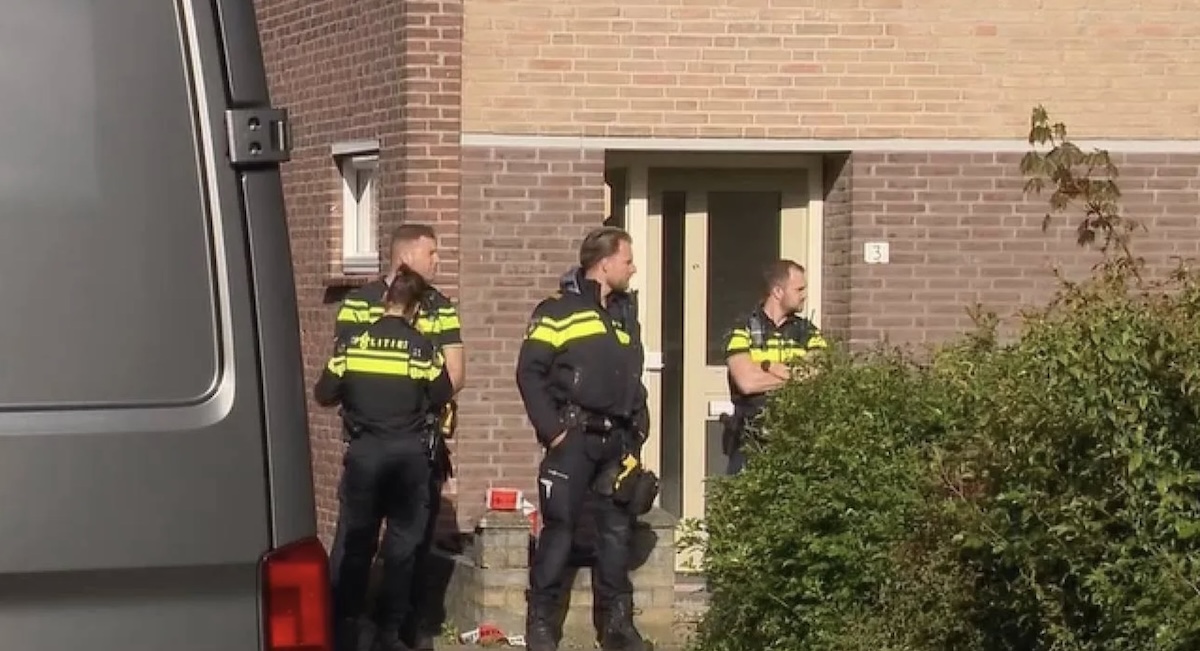 Three firefighters in black and yellow turnout gear stand outside a brick house entrance beside a hedge and a vehicle to the left