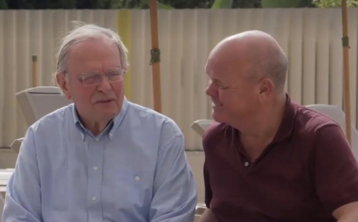 Two men sit together outdoors one in a light blue shirt and the other in a burgundy polo sharing a friendly conversation