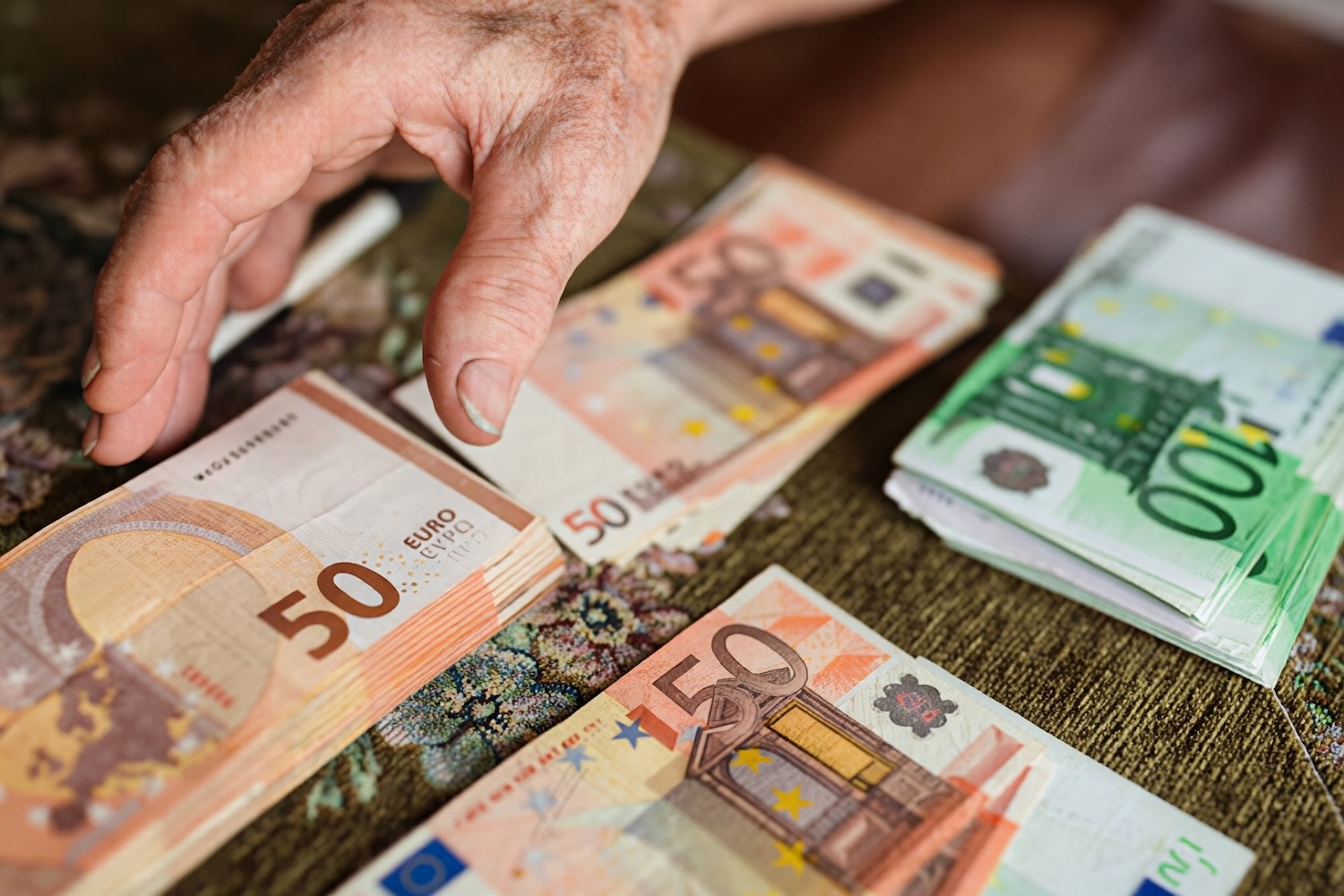 Person's weathered hand counting euro banknotes on a table with 50€ and 100€ bills visible