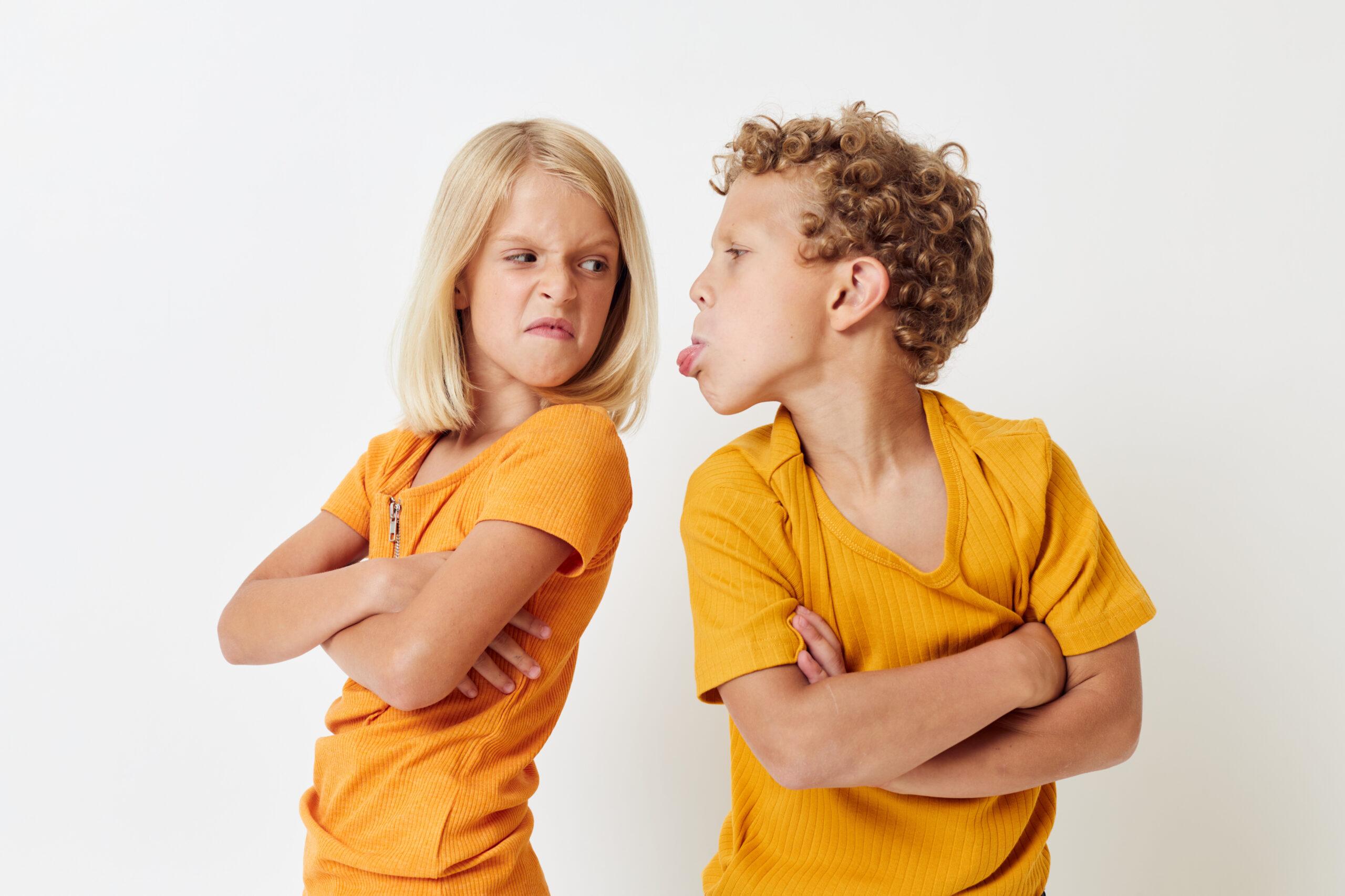 Two children facing each other with arms crossed making goofy faces in orange shirts against a white background