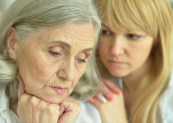 Older woman with hands under her chin eyes closed as a younger woman offers supportive presence from behind