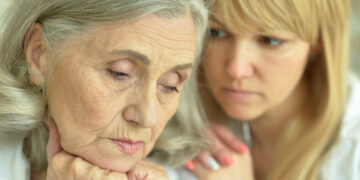 Older woman with hands under her chin, eyes closed, as a younger woman offers supportive presence from behind.
