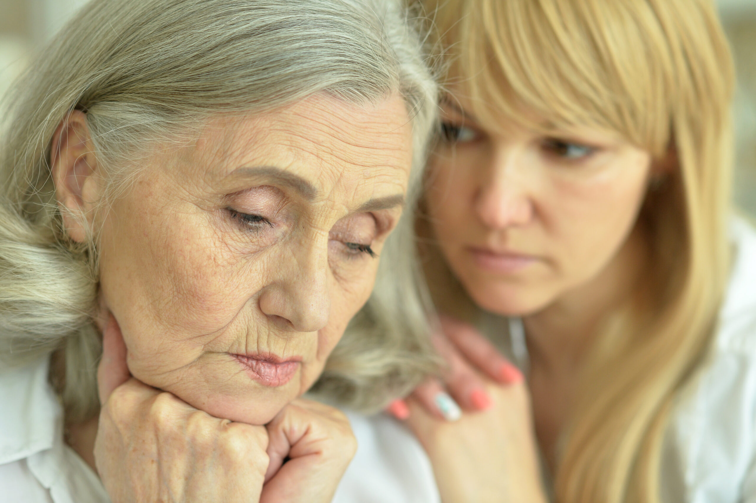 Older woman with hands under her chin eyes closed as a younger woman offers supportive presence from behind