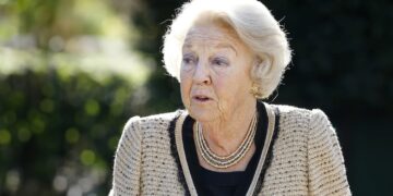 Elderly woman with white hair wearing a beige tweed jacket and layered necklace outdoors.