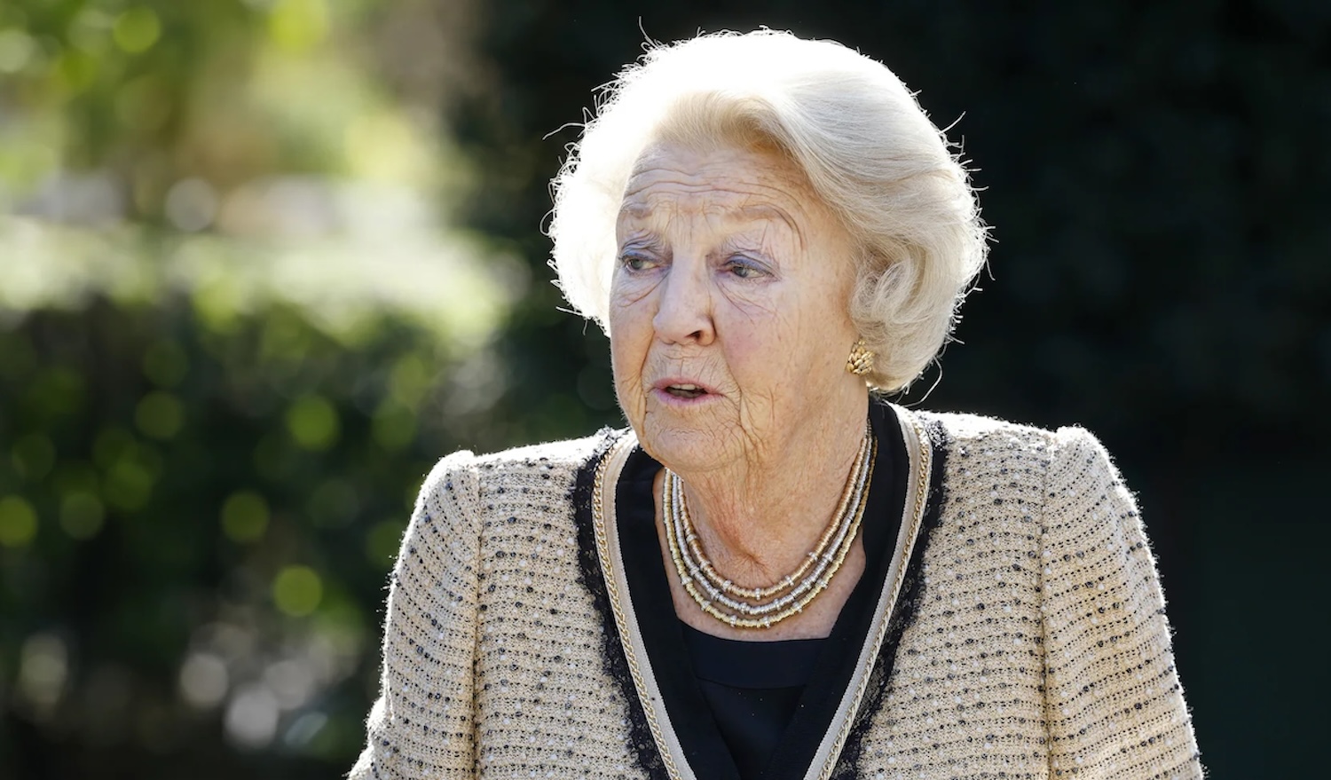 Elderly woman with white hair wearing a beige tweed jacket and layered necklace outdoors