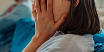 Woman covering her face with both hands while seated in a waiting area; man blurred in the background.