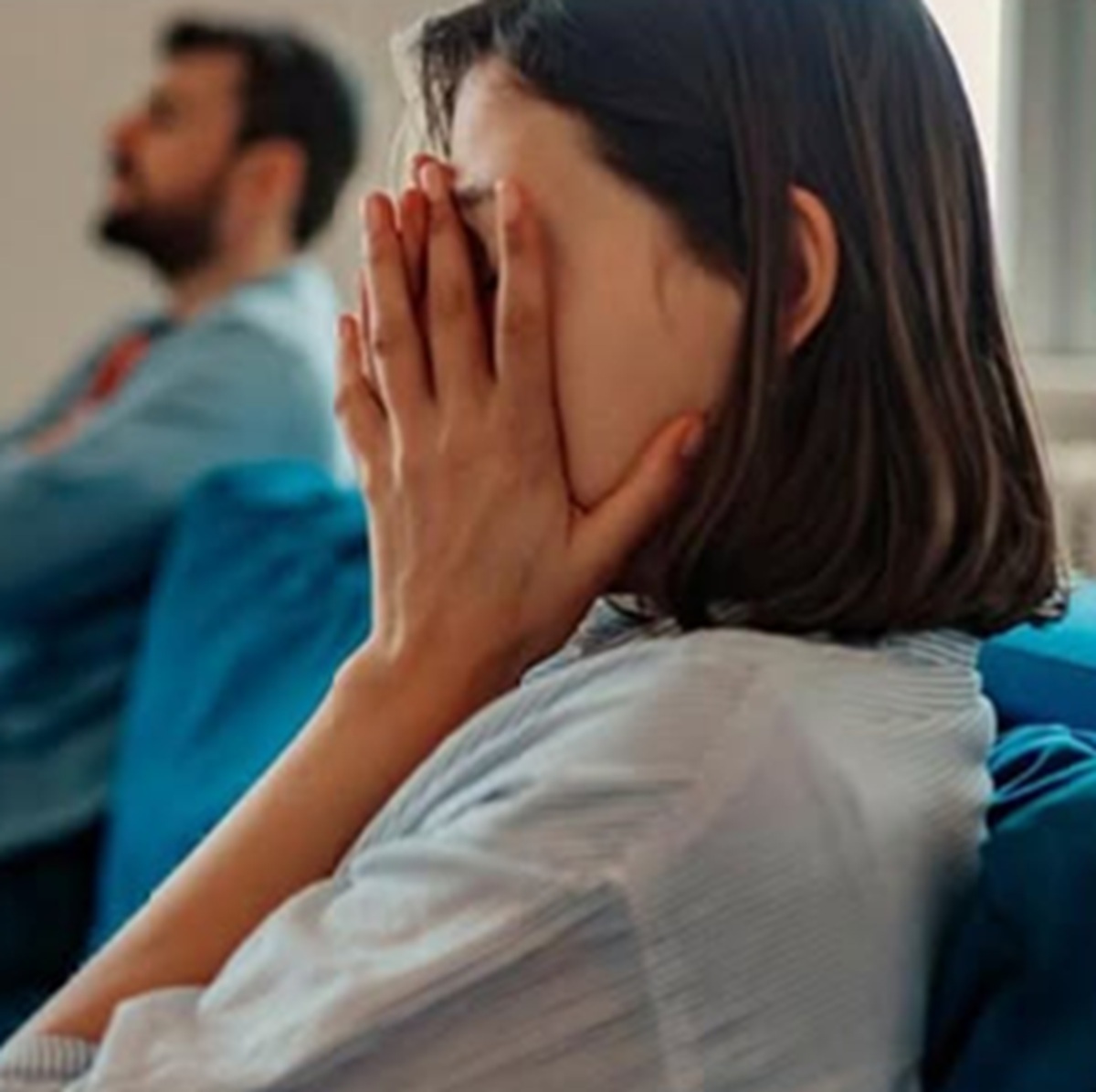 Woman covering her face with both hands while seated in a waiting area man blurred in the background
