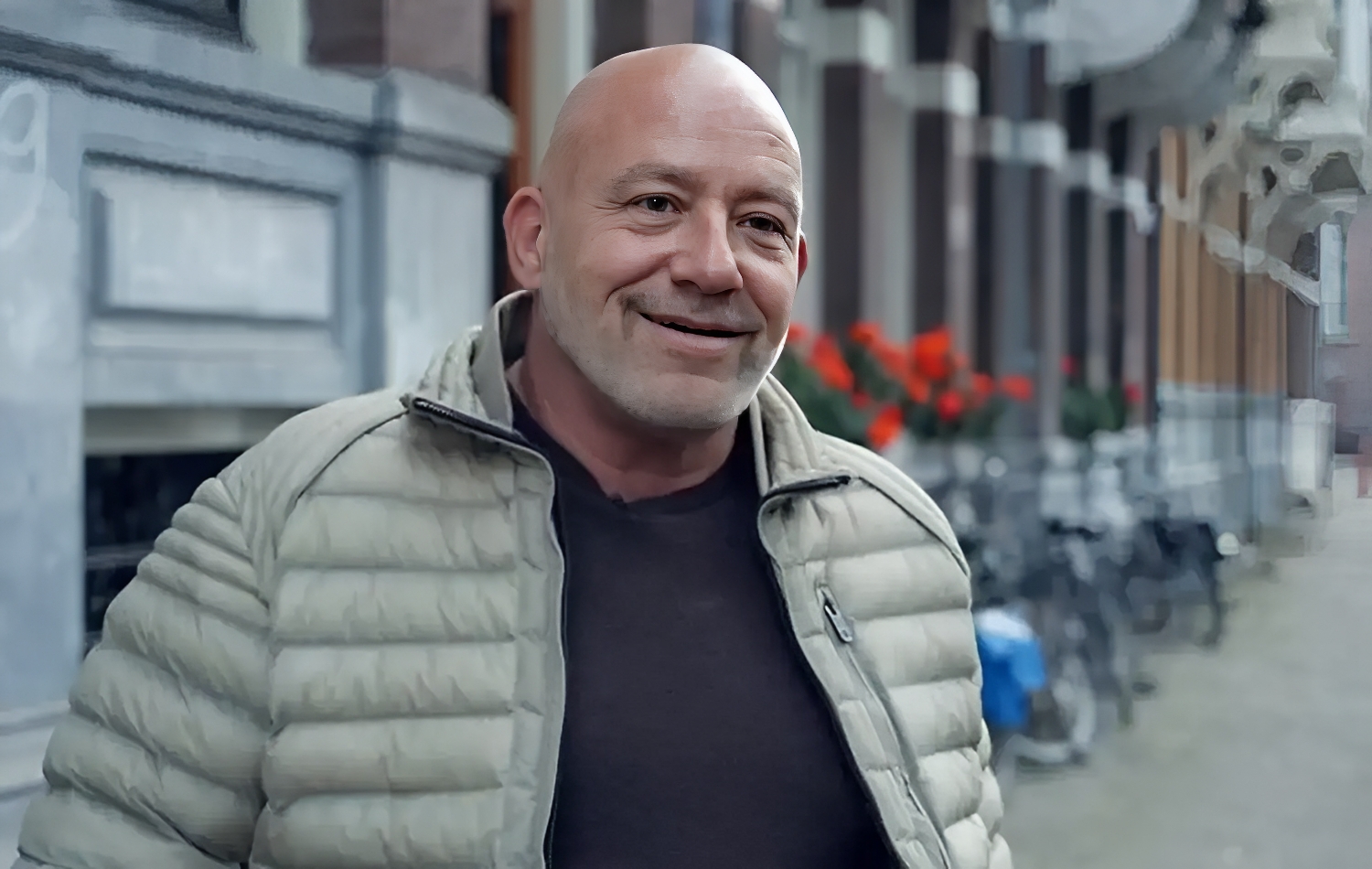 Smiling bald man in a light puffer jacket on a city sidewalk with red flowers in the background 