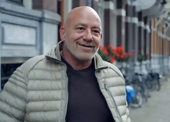 Smiling bald man in a light gray puffer jacket standing on a city street with red flowers in the background