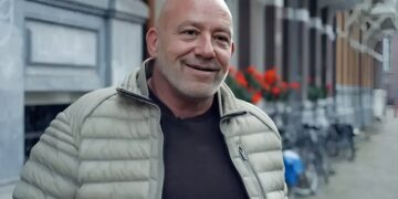 Smiling bald man in a light gray puffer jacket standing on a city street with red flowers in the background