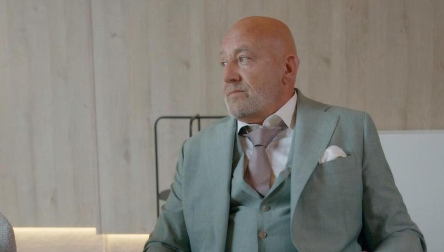 Older man in a light green suit with vest white shirt and pink tie seated indoors and looking to his left