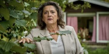 Woman in a light blazer standing outside near a house with pink curtains, looking concerned or surprised.
