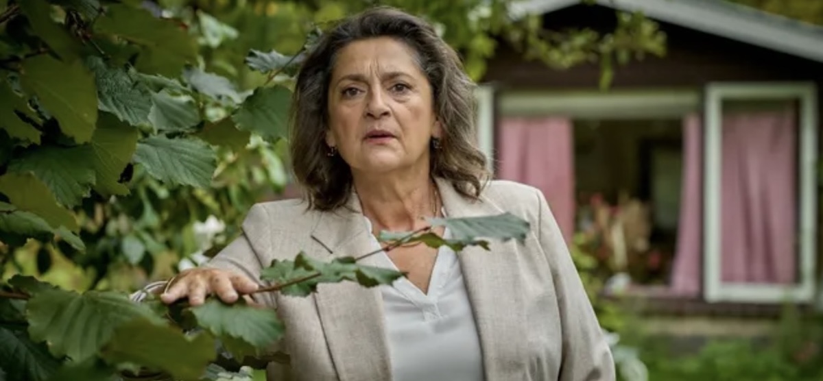 Woman in a light blazer standing outside near a house with pink curtains looking concerned or surprised