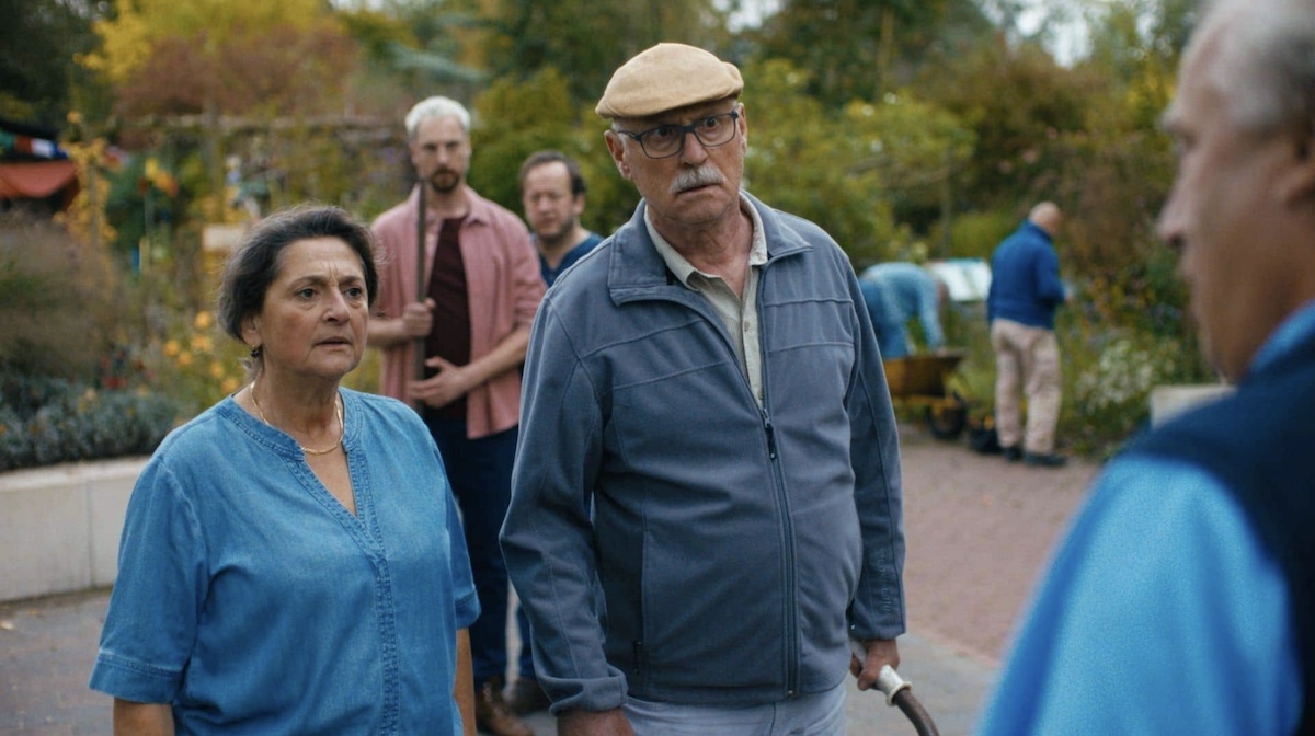 An elderly man in a tan cap and blue gray jacket stands beside an older woman in a blue blouse outdoors in an autumn garden