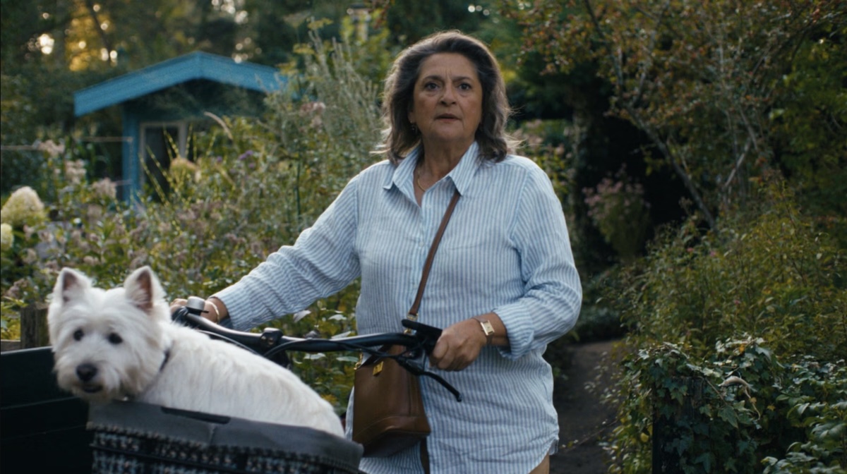 Older woman in a light blue striped shirt rides a bicycle with a white fluffy dog in a garden setting