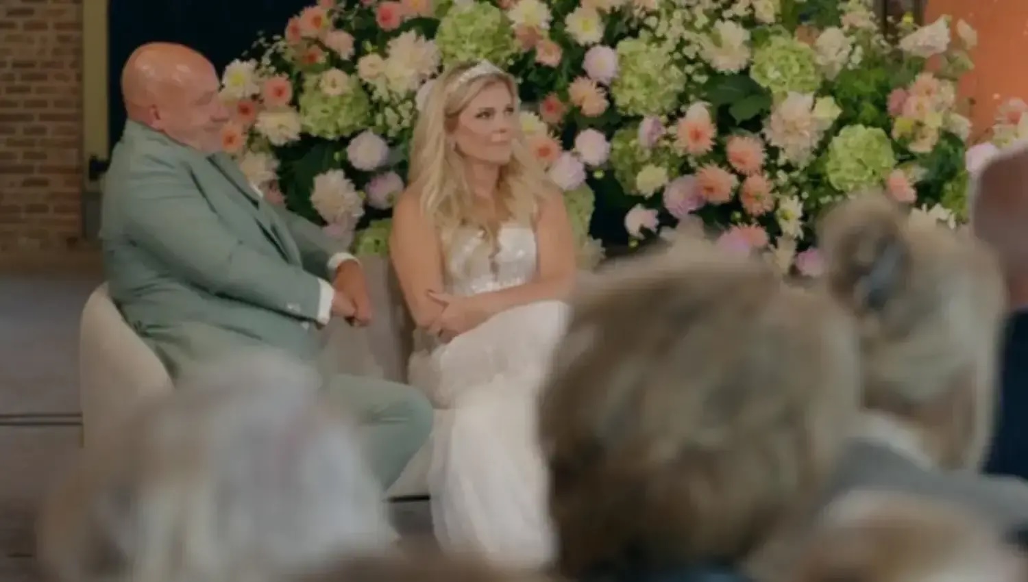 A bride in a white dress sits beside a man in a light suit on a stage with a wall of pink and white flowers behind them during a ceremony or reception